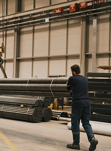 A man in work clothes pulling or moving bundled steel pipes in a warehouse or industrial setting with large shelves and concrete floor.