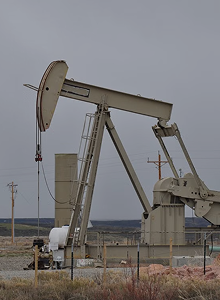 An oil pumpjack in a fenced area with a cloudy sky and utility poles in the background.