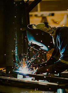 Person wearing a protective welding helmet and gloves welding metal with bright sparks flying around in a workshop.