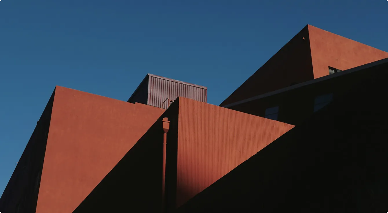 Angular red and brown building structures with sharp edges and shadow patterns against a clear blue sky.